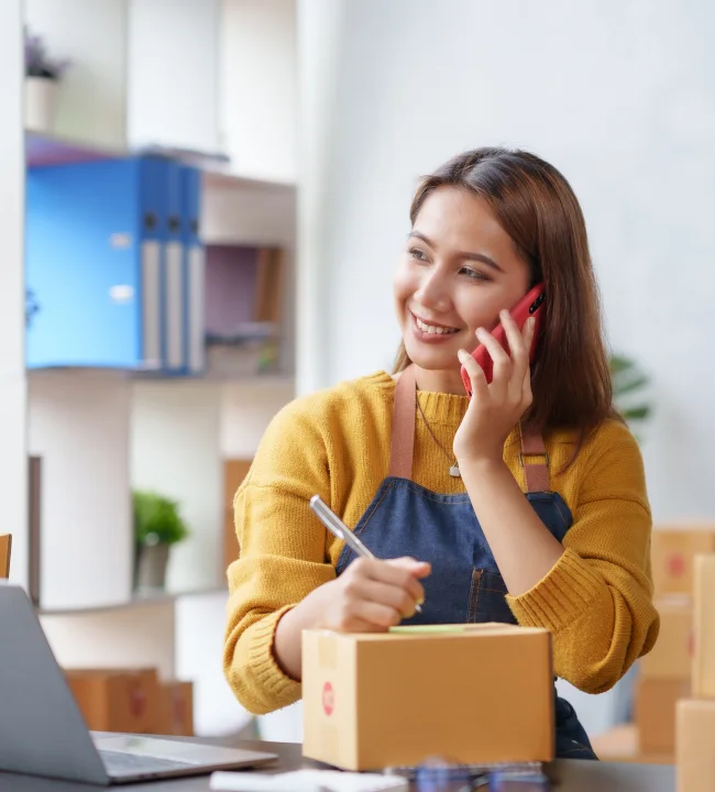 Online seller taking a customer call at her desk beside a laptop and packed parcels